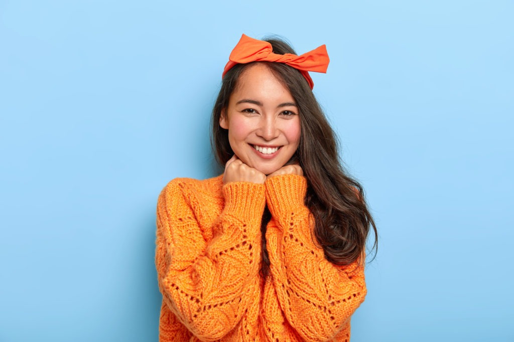 Portrait of glad brunette millennial female keeps both hands under chin, smiles pleasantly, wears orange bow headband, warm orange knitted sweater, expresses sincere feelings. Face expressions orthodontist owensboro ky