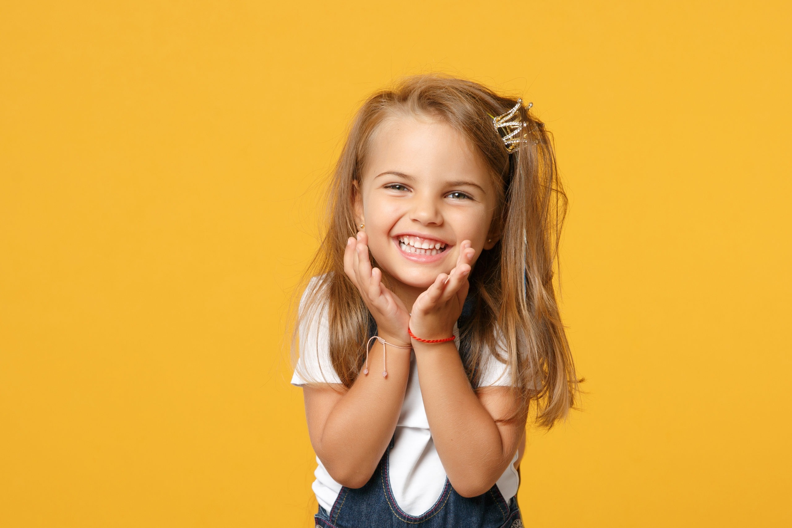 Smiling girl with long hair in a white shirt and denim overalls, showcasing her teeth against a bright yellow background, representing the joy of orthodontic treatment for children.