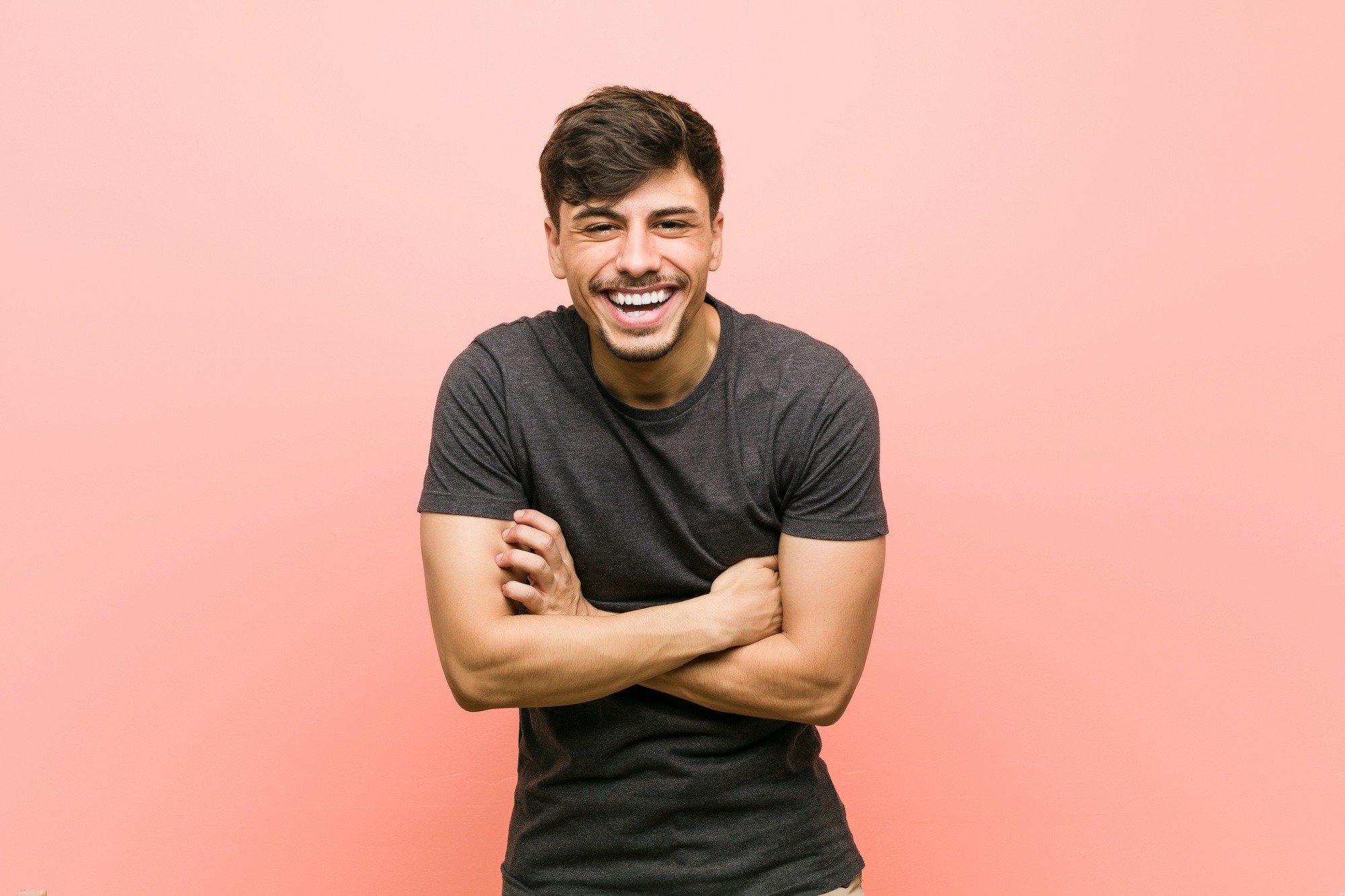 Smiling young man in a gray t-shirt, arms crossed, against a pink background, representing confidence and satisfaction with orthodontic treatment options.